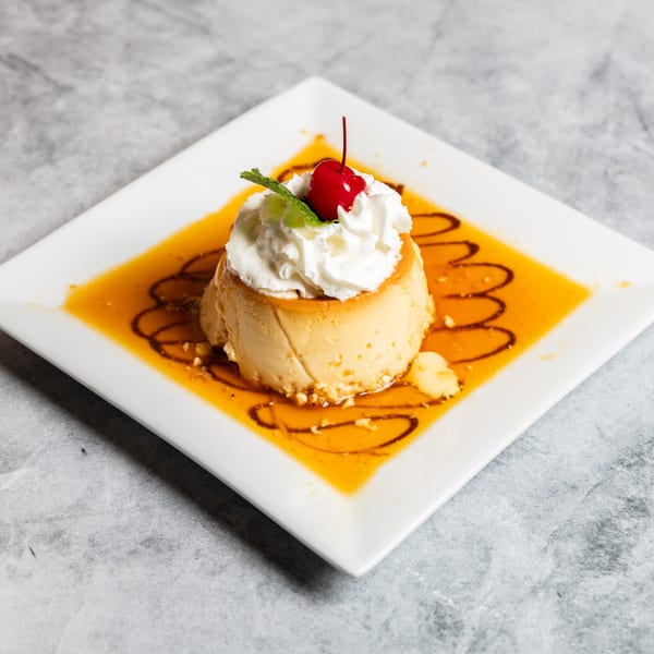 Creamy flan dessert topped with whipped cream, a cherry, and mint, surrounded by caramel sauce.