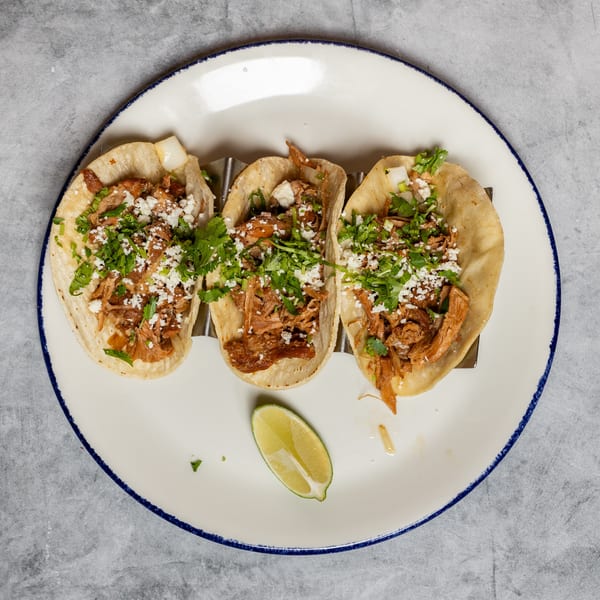 Three tacos filled with shredded meat, topped with cilantro, crumbled cheese, and a lime.
