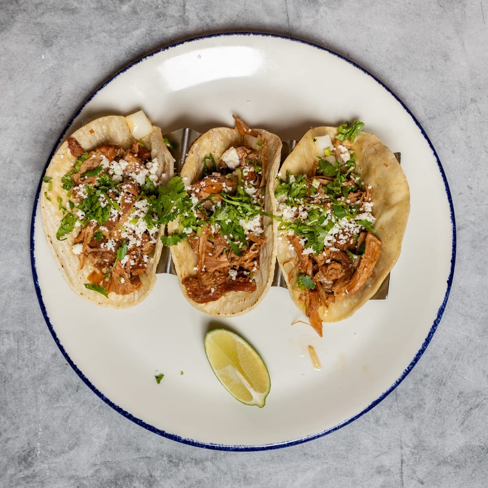 Three tacos filled with shredded meat, topped with cilantro, crumbled cheese, and a lime.