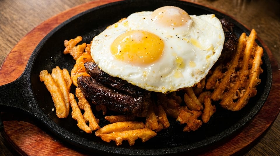 Sizzling steak and waffle fries topped with two fried eggs in a black skillet.