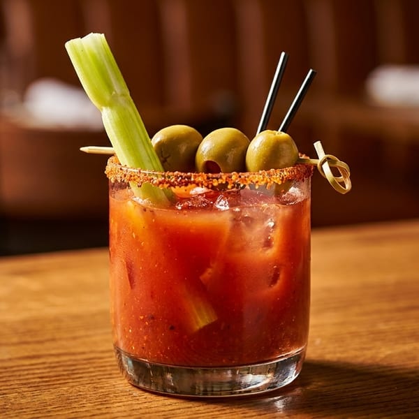 Bloody Mary cocktail garnished with celery, olives, and a spiced rim on a wooden bar.