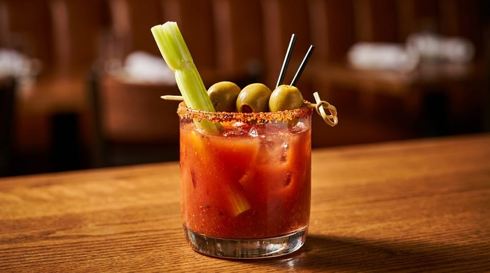 Bloody Mary cocktail garnished with celery, olives, and a spiced rim on a wooden bar.