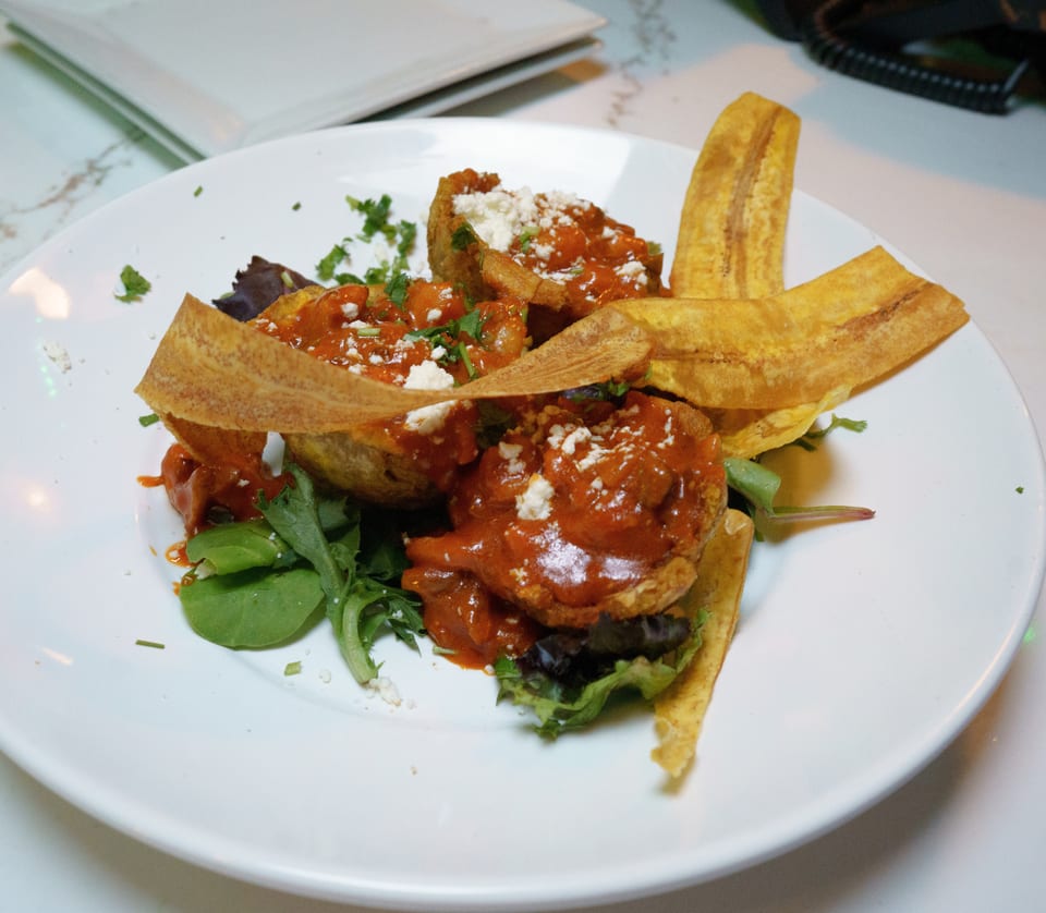 A white plate holds fried plantain cups filled with red sauce, cheese, and herbs, with plantain chips.