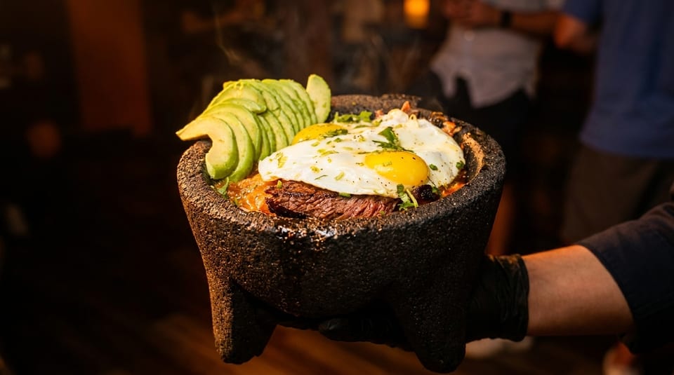 Steaming molcajete bowl with steak, fried eggs, avocado slices, and cilantro, held by a gloved hand.