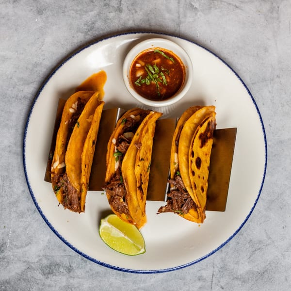 Three birria tacos in a holder with dipping sauce and a lime on a white plate.