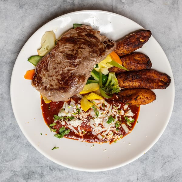 Grilled steak, mixed vegetables, fried plantains, and red sauce with cheese on a white plate.
