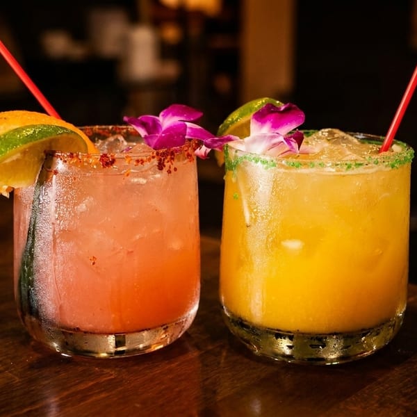 Two colorful cocktails, one pink and one yellow, garnished with fruit and flowers on a wooden table.