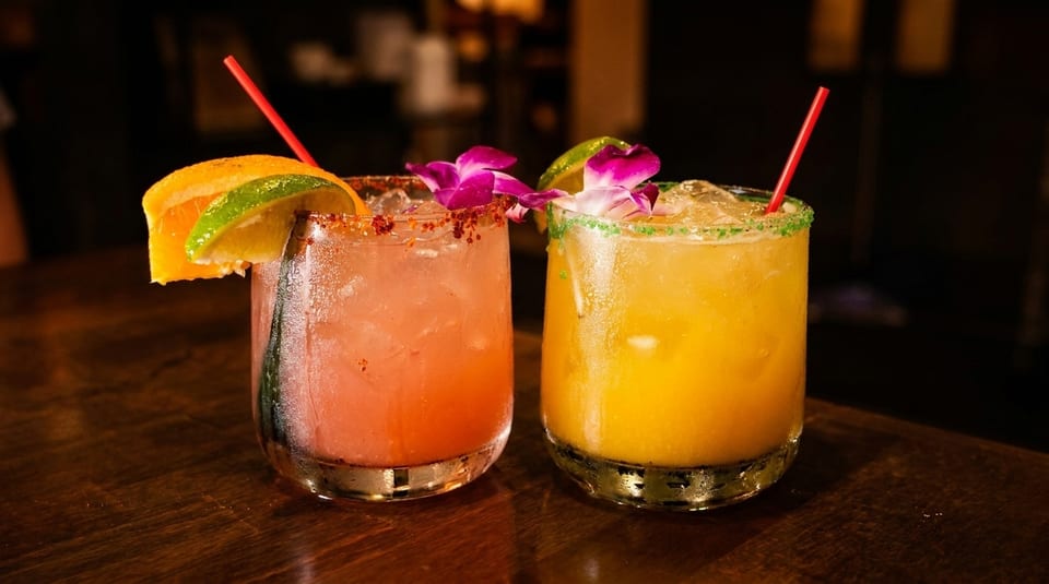Two colorful cocktails, one pink and one yellow, garnished with fruit and flowers on a wooden table.