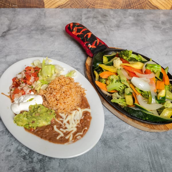 Plate of Mexican food with rice, beans, guacamole, and pico de gallo next to a sizzling skillet of mixed vegetables.