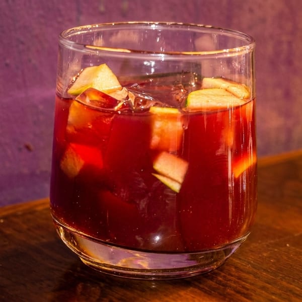A glass of red sangria with apple chunks and ice sits on a wooden table.