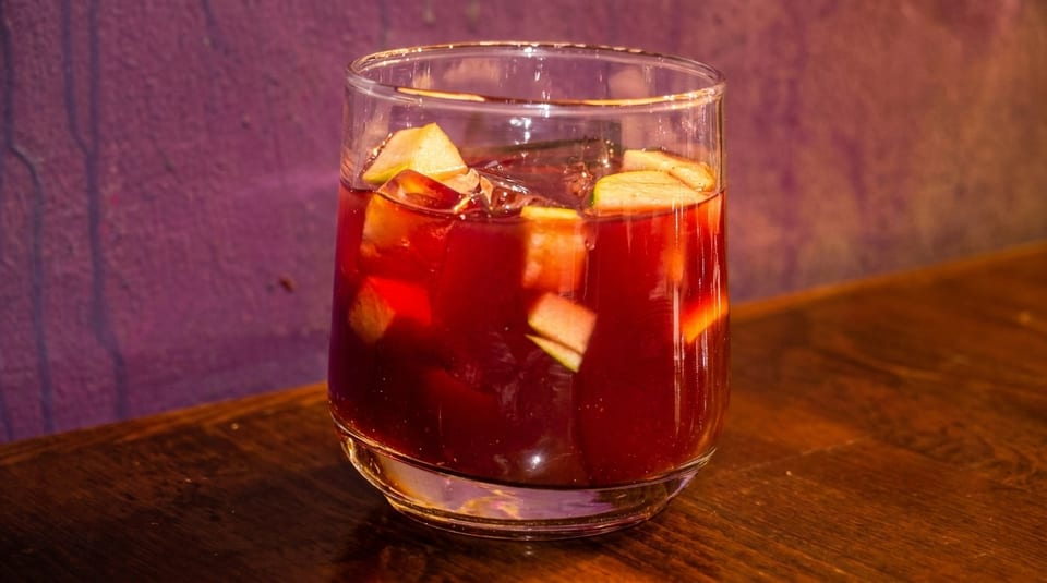 A glass of red sangria with apple chunks and ice sits on a wooden table.