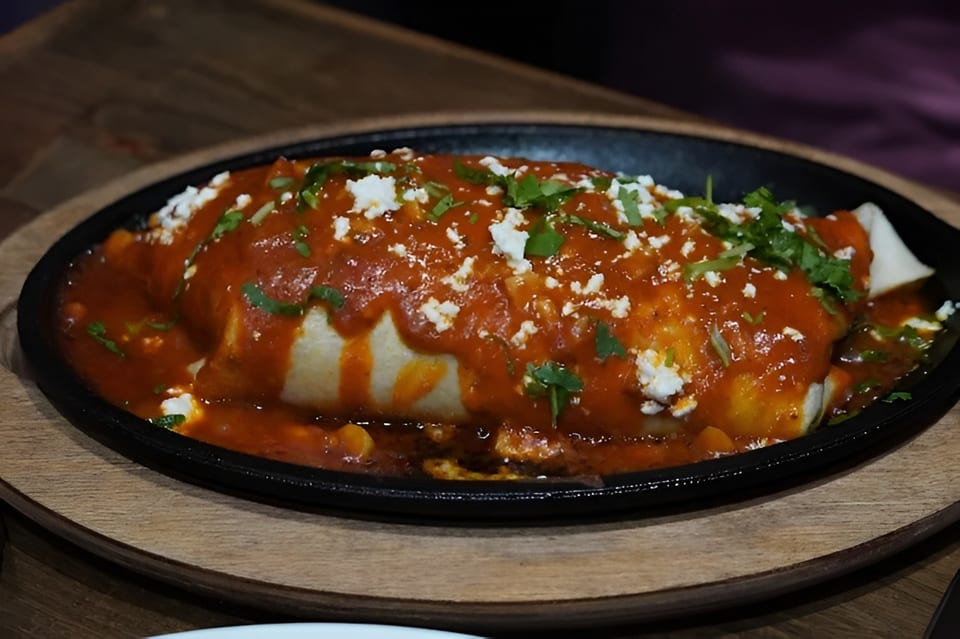 A sizzling burrito covered in red sauce, crumbled white cheese, and cilantro, served on a black platter.